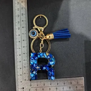 Gedetailleerd uitzicht van blauwe letter R sleutelhanger met boze oog bedel en kwast op liniaal.