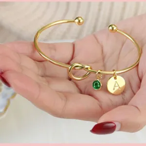 Bracciale rigido dorato regolabile con nodo, ciondolo lettera A e pietra portafortuna verde.