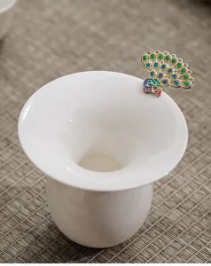 Close-up detail of white ceramic tea strainer with elegant peacock metal handle accessory.