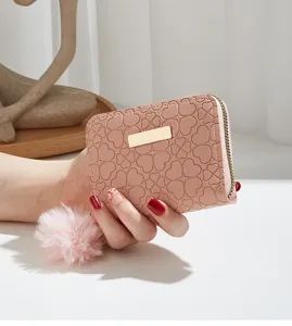 Pink wallet detail view showing heart embossed pattern, golden metal plate and fluffy pom-pom charm.