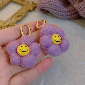 Detail shot of purple fluffy flower earrings with cute yellow smiley face and gold-tone huggie hoops.