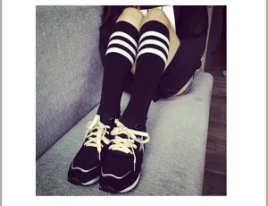 Detail view of black knee high socks with three white stripes worn with casual sneakers on sofa.