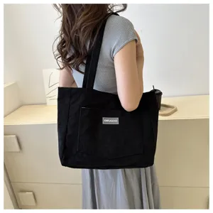 Close-up of a black corduroy tote bag worn on shoulder, showing textured fabric and front pocket.