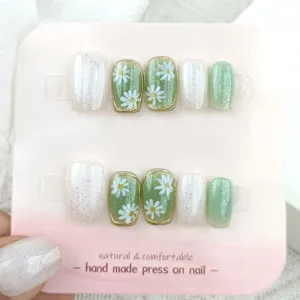 Detalle de uñas postizas hechas a mano con diseño floral de margaritas verdes y brillo.
