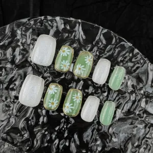 Primer plano detallado de uñas postizas verdes con margaritas, purpurina y bordes dorados.