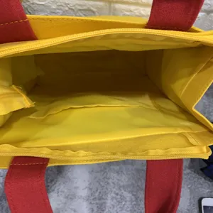 Vista interior detallada del bolso de lona amarillo con asas rojas y bolsillo con cremallera.