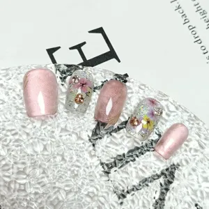 Uñas postizas hechas a mano cortas con efecto ojo de gato rosa, flores secas y pedrería brillante.