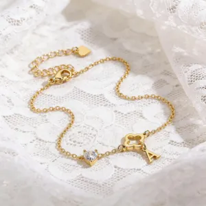 Detailaufnahme eines goldenen Kleeblatt- und Buchstaben-Armbands auf weißer zarter Spitze.