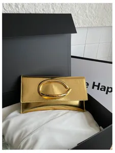 Gouden metallische envelop clutch tas met opvallend sculpturaal C-vormig metalen handvatdetail.