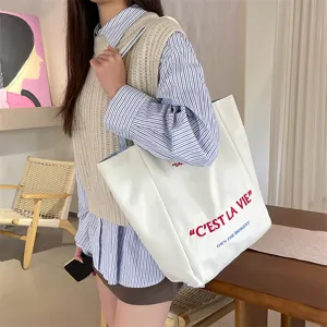 Close-up of a stylish white canvas tote bag with 'C'EST LA VIE' embroidery on a model.