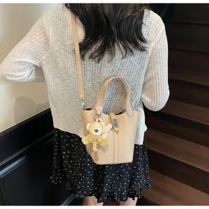 Detalles del bolso cubo beige con patrón litchi, colgante de oso y correa ajustable.