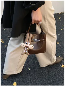 Dettaglio di una borsa a secchiello in pelle marrone con chiusura in metallo e ciondolo peluche.