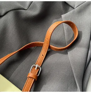 Primer plano de correa de bolso de cuero marrón con hebilla de metal sobre tela gris texturizada.