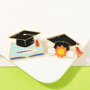 Nahaufnahme von Abschluss-Emailleschmuck mit Kappe, Buch und Sonnenblumen-Motivdetails.