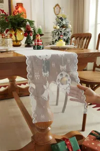Camino de mesa navideño de encaje blanco con motivos de copos y campanas para decoración festiva.