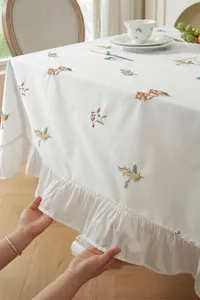 Close-up detail of white cotton tablecloth with floral embroidery and ruffled hem.