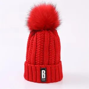 Red winter knitted beanie hat with fluffy pom pom and trendy letter patch detail.