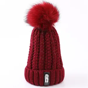 Red winter knitted beanie hat with fluffy faux fur pom pom and stylish letter label detail.