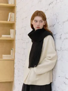 Detailed shot of a classic black knitted scarf worn by a model against a textured white wall.