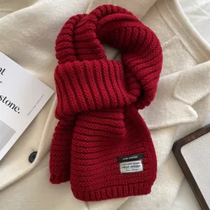 Close-up detail of a cozy red chunky knit scarf with brand label on a soft white coat background.