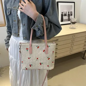 Close-up lifestyle view of the woven straw tote bag featuring cute cherry embroidery and pink straps.