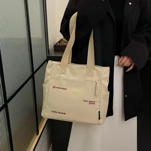 Canvas tote bag detail view, showing beige fabric, red embroidery text, and side pocket design.
