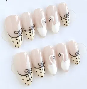 Detailed close-up of almond press-on nails with nude base, black bow, polka dots and swan art.