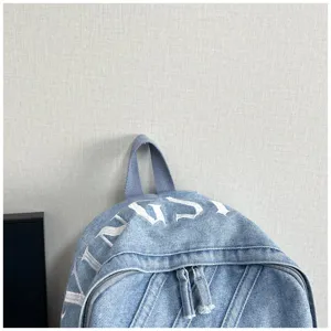 Denim backpack detail showing washed fabric texture, white embroidery letters and sturdy top handle.