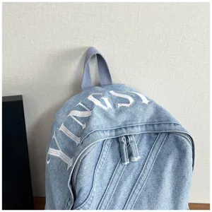 Detail view of the denim backpack top handle, dual zippers, and white embroidery design.