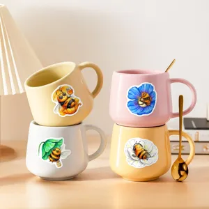 Close-up of cute ceramic coffee mugs with watercolor bee illustrations and gold spoon on desk.