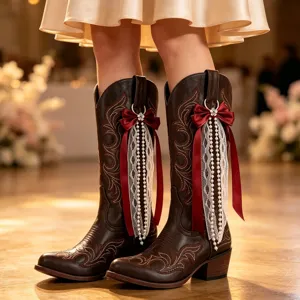 Close-up of brown western cowboy boots with red bow and pearl lace fringe charms.