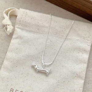 Gros plan détail du collier pendentif chien teckel argenté sur sachet en tissu, style épuré.