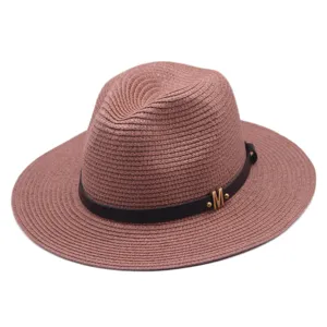 Detailaufnahme des rosafarbenen Stroh-Fedora-Hutes mit schwarzem Gürtel und goldenem M.