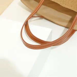Detailed view of women woven straw tote bag brown leather handles with white stitching design.