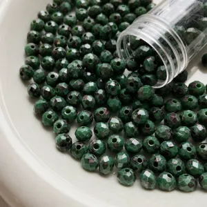 Macro detail of green faceted fire polished glass beads spilling from tube on white plate.