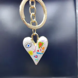 Close-up gold keychain with enamel heart pendant, colorful artistic eye and flower doodle details.