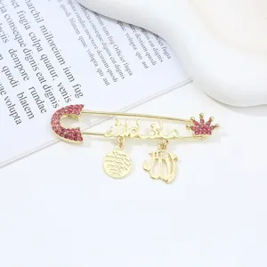 Gold-tone safety pin brooch with pink rhinestones, crown charm, and Arabic calligraphy details.