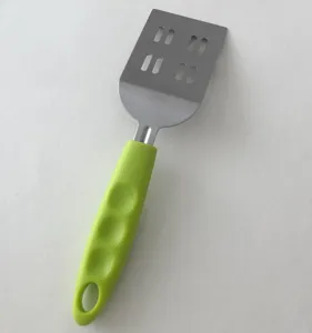 Stainless steel spatula detail showing slotted metal blade and ergonomic green handle.