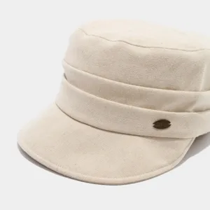 Primer plano detallado de la gorra plana beige que destaca el elegante emblema de la marca metálica.
