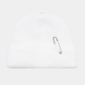 Detailopname van een witte gebreide muts met een metalen veiligheidsspeld als accent.