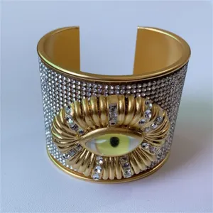 Bracciale rigido dorato largo con strass brillanti e un dettaglio di occhio surreale.