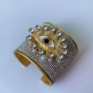 Bracciale polsino largo dorato con motivo malocchio, strass e perle alla moda.