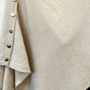 Close-up detail of beige knitted poncho featuring soft fabric texture and metal buttons.