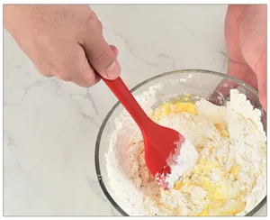 Primo piano dettagliato di una spatola in silicone rosso che mescola la farina in una ciotola.