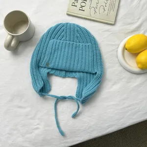 Primer plano del gorro de punto azul con orejeras y lazos sobre tela blanca con accesorios.