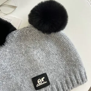 Grey knit beanie detail showing soft wool texture, black pom poms, and logo patch.