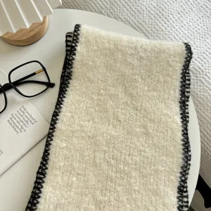 Close-up of cozy cream knitted scarf with black edge stitching on white table with lifestyle props.