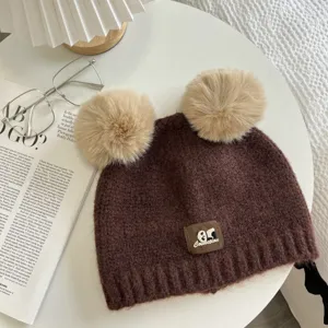 Close-up detail of a brown knitted beanie hat featuring dual fluffy pom-poms and front label.