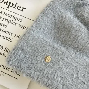 Grey fuzzy knit beanie detail close-up, soft rabbit hair blend fabric with gold metal logo tag.