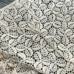 Primo piano di tessuto in pizzo crochet beige con delicato motivo floreale e frange di conchiglie.